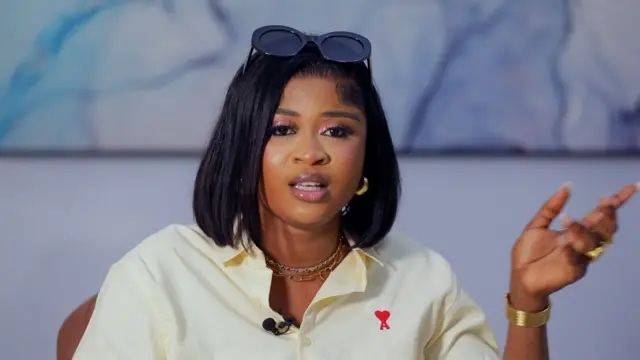 Imisioluwa Ayanwale to se ipo kinni nile Big Brother Naija fun ọdun 2025 joko lori aga kan, o na ọwọ osi soke bii ẹni to n ta ika, o fi irun wiigi dudu sori, o si fi igo akọ soke sori irun rẹ, o wọ asọ shaat alawọ miliki, o ọ sokoto alawọ buluu, to si lo bẹliti sori sokoto naa.