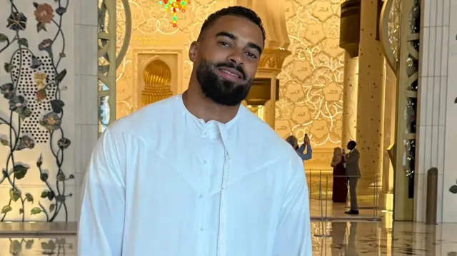 Latif Ayodele, con camisa blanca, sonríe a la cámara. Está de pie dentro de un edificio ornamentado.