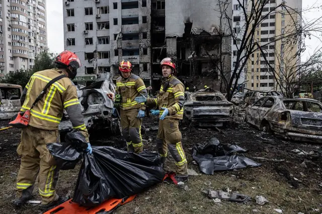 На передньому плані троє рятувальників у захисному обладнанні та гумових рукавичках стоять біля тіла, що лежить у чорному пакеті на ношах.
За ними – згорілі автомобілі та сліди пожежі, на задньому плані — пошкоджена багатоповерхівка з вибитими вікнами, знесеною повністю стіною, через яку проглядаються спалені квартири на щонайменше трьох поверхах.