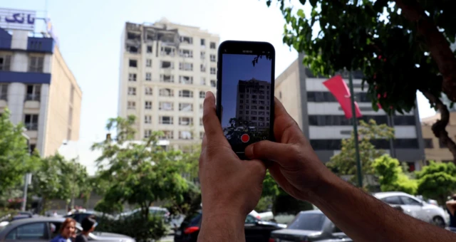 Tahran'da beyaz renkli bir binanın çatı katı vurulmuş, sokaktan bir kişi cep telefonu ile bu vurulan binayı fotoğraflıyor.