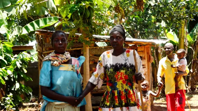 Deux femmes se tenant par la main, la femme de droite cherchant à être guidée (devant) et un père avec ses enfants (derrière) à l'extérieur, au soleil.