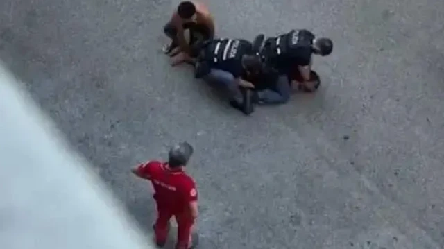 عکسی از بالا گرفته شده که سه پلیس مردی را روی زمین خوابانده و روی او نشستهاند و در حال بازداشت او هستند، یک پلیس هم در کنار ایستاده در حال تماشاست
