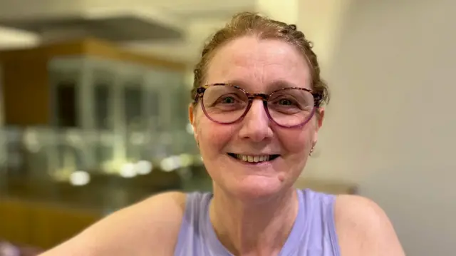 Kate is wearing gym clothes and has her hair tied in a ponytail. She is wearing pink-rimmed glasses.