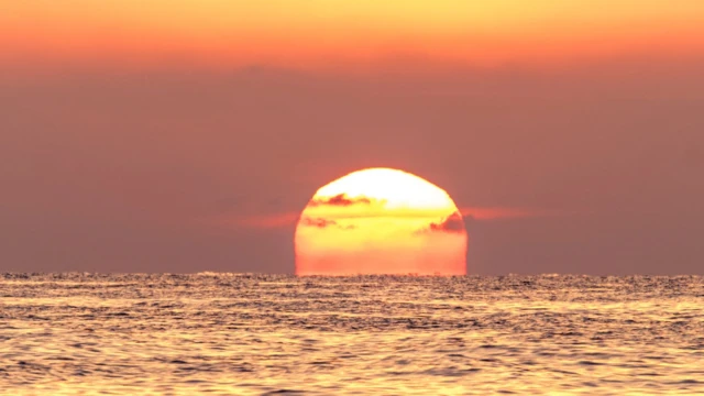 Imagem do sol vermelho sobre o mar e com um céu laranja.