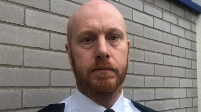 John Massey: la cabeza y los hombros de un hombre calvo con barba rojiza de pie frente a una pared de ladrillos de color marrón pálido.