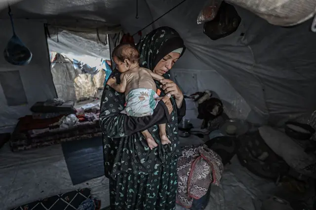 الأم ترتدي حجاباً وتحمل طفلها الذي يبدو عليه الهزال بسبب الجوع، داخل خيمة.