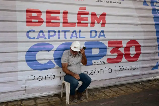 Operário sentado em frente a painel anunciando COP30 em Belém