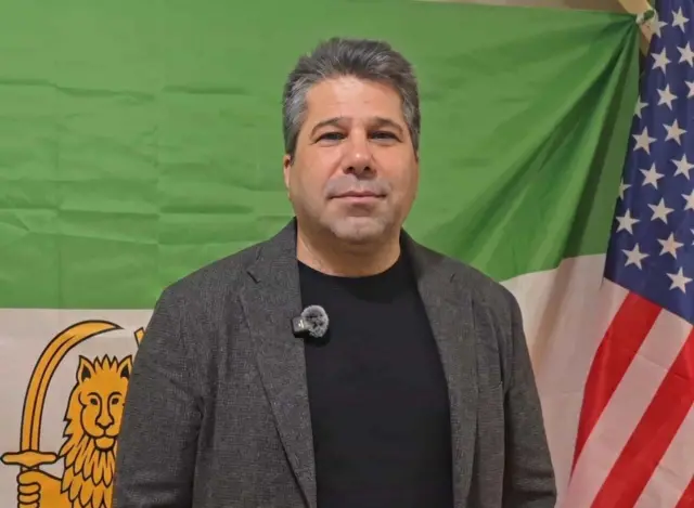 عکس مسعود طاهری در حالی که پشت سرش پرچم شیر و خورشید و پرچم آمریکا دیده می‌شود.