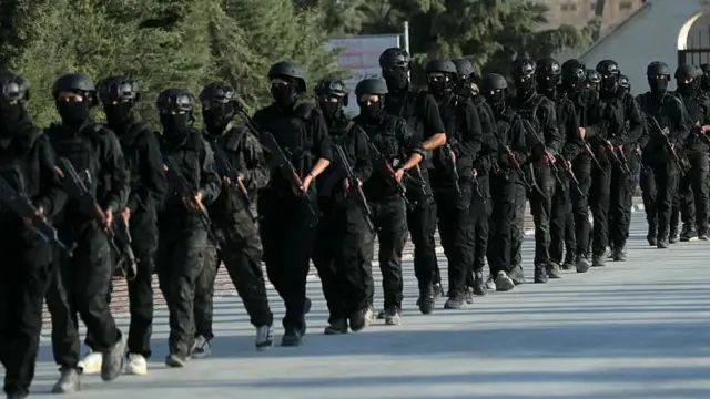 Siyah kamuflajlı ve siyah miğferli askerler ellerinde tüfeklerle yürüyor. Fotoğraf gündüz çekilmiş. Yerde asfalt, arkada ise yeşil ağaçlar görünüyor.