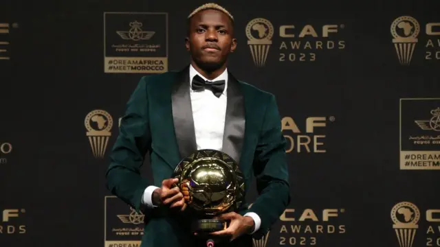 Victor Osimhen en smoking vert et nœud papillon noir pose avec son trophée de Joueur africain de l'année devant les panneaux faisant la promotion des Caf Awards 2023