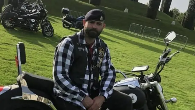 Anthony Frith está sentado en una motocicleta en el Arboreto Nacional del Recuerdo. Lleva una boina, una camisa a cuadros blancos y negros y una chaqueta sin mangas con medallas. Al fondo se ve el Monumento a las Fuerzas Armadas, dos motocicletas y varios otros motociclistas.