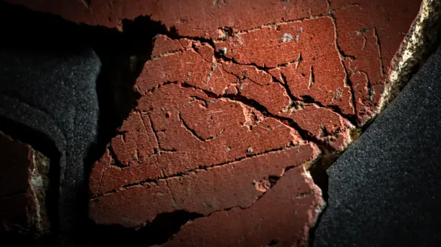 Un primer plano de un trozo de yeso rojo, formado por varios trozos más pequeños, con las letras F E C I T en mayúsculas claramente visibles con una línea dibujada en forma de cuadrado alrededor de tres lados de la palabra. Falta el lado izquierdo del yeso, donde debería estar el nombre de la persona o grupo que realizó la obra de arte.