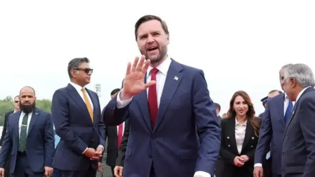 Visi Perezida w'Amerika JD Vance apepera n'ikiganza. Yambaye ikositimu y'ubururu, ishati y'umweru na karuvati itukura. Inyuma ye n'iruhande rwe hari abantu benshi.