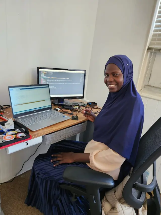 Aminata Sarr en hijab bleu devant deux écrans d'ordinateurs allumés, elle sourit à la caméra.