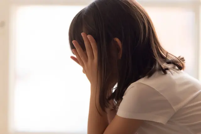 Menina branca de cabelos castanhos no ombro, vista de perfil, esconde o rosto nas mãos