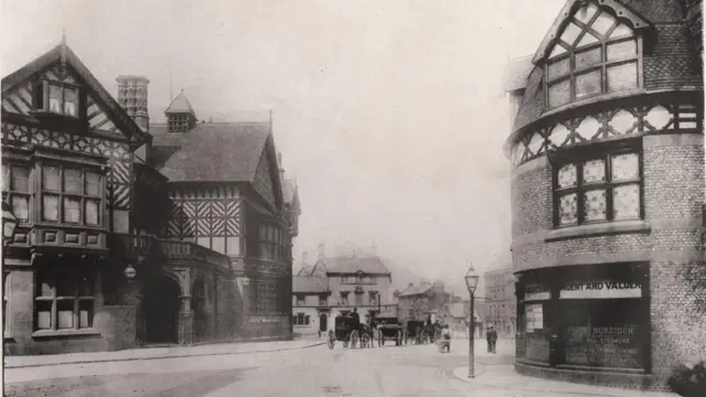 Fotografía de época en blanco y negro de la Plaza del Mercado Viejo de Altrincham, procedente del archivo del Ayuntamiento de Trafford. Dos edificios con fachadas en blanco y negro, con techo de paja, se encuentran a ambos lados de la calle. Al final de la calle se ven tres caballos y carretas.
