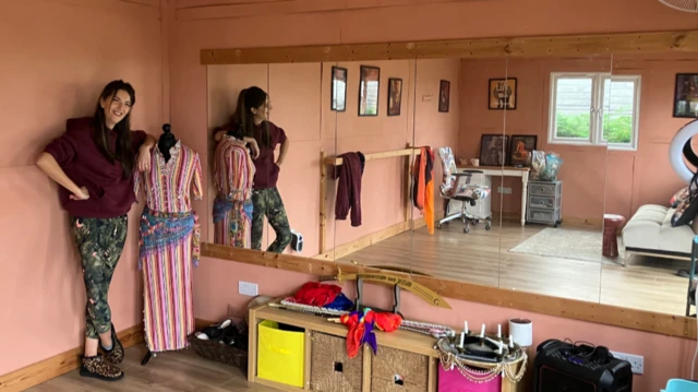 Una mujer con un top burdeos y leggings florales se apoya en un maniquí que viste un vestido de rayas de colores. Hay grandes espejos a lo largo de una pared de la sala, donde se pueden ver una pista de baile, un escritorio y un sofá.
