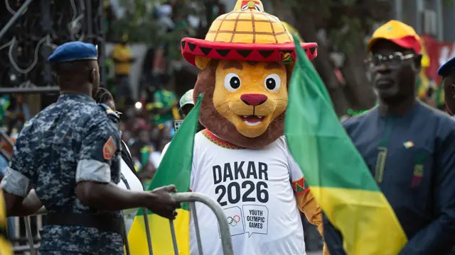 Une mascotte de lion à l'air sympathique, au visage brun doré, aux grands yeux expressifs et au large sourire, porte un t-shirt blanc où l'on peut lire « Dakar 2026 » au-dessus du logo officiel des Jeux Olympiques de la Jeunesse. Elle arbore également un chapeau conique surdimensionné et distinctif, orné de motifs jaunes et orange et d'un bord rouge. À gauche, un agent de sécurité en uniforme de camouflage bleu et béret bleu, dos à la caméra, gère une barrière métallique de sécurité. Flou sur la droite, un homme en tunique traditionnelle sombre et casquette jaune et rouge observe la scène en tenant un drapeau sénégalais partiellement plié. À l'arrière-plan, une foule dense et floue de spectateurs se dessine.