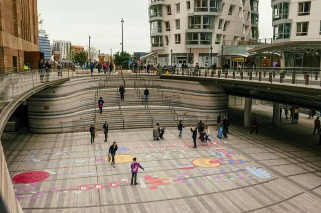 Yüksek binaların ortasında bir meydan, çocuklar renkli çizimlerin etrafında oynuyor.