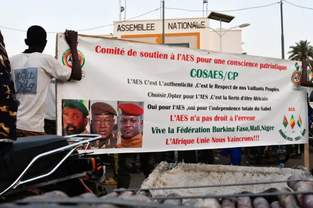 Une banderole sur laquelle se trouvent les trois images des trois militaires qui ont formé l'AES, avec derrière un bâtiment sur lequel est écrit "Assemblée nationale".