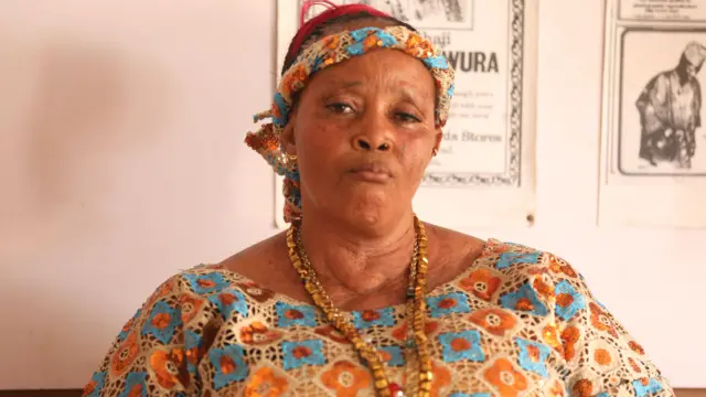 Kabirat Ayinla Omowura wọ aṣọ léèsì alawọ buluu ati goolu, o we gele asọ leesi naa, o si fi ilẹkẹ meji sọrun, aworan meji to jẹ ti baba rẹ, tii se gbajumọ akọrin Apala nigba aye rẹ, Ayinla Omowura, wa lara ogiri ti obinrin naa fẹyin ti.