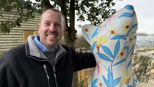 Ian Ridpath: un hombre sonriente con una chaqueta oscura, de pie junto a una colorida escultura de una rata. La obra presenta flores azules y amarillas y mira hacia arriba.