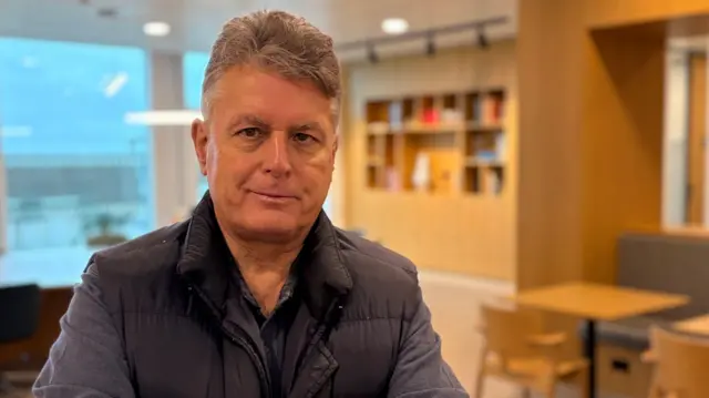 A head and shoulders image of JP Allard in a modern-looking office area. He is wearing a dark gilet over a long sleeved casual blue shirt, which is open at the collar. He has relatively short, light coloured hair. Behind him, though out of focus in the picture, are chairs, tables and a book case.