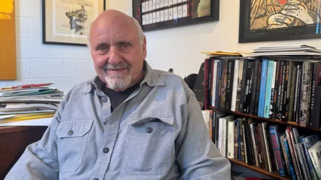 Um homem vestindo uma camisa cinza sentado em frente a uma estante de livros