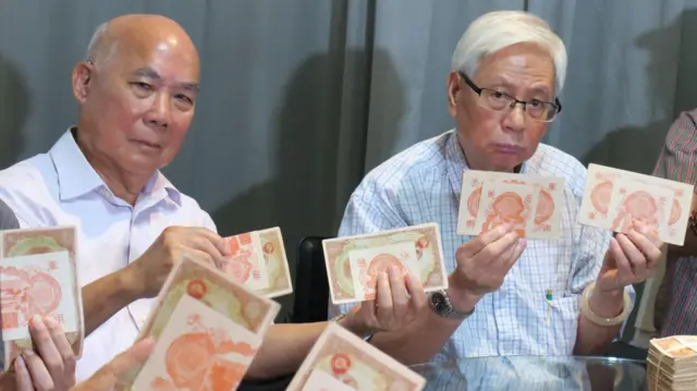身穿白色配蓝色格仔花纹衬衣的莫国威（右）与身穿纯白色衬衣的刘文（左）举起日占香港军票展示（BBC中文图片）