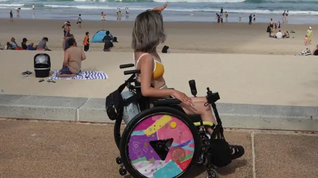 یوه ښځه چې لنډ ویښتان لري، په ویلچېیر کې د سمندر پر غاړه ناسته ده او هماغې خوا ته ګوري.