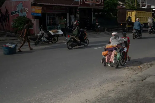Seorang anak mendorong kursi roda ibunya yang cacat, mereka memakai masker wajah dan pelindung plastik di tengah pandemi virus corona COVID-19, menjual makanan ringan di sepanjang jalan di Pekanbaru, Riay