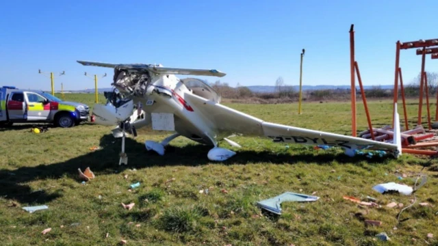 A crashed light aeroplane on an airfield, surrounded by debris, with bright orange technical structures beside it. There is an emergency vehicle behind the plane and the nose is completely destroyed. It is a sunny day with a blue sky.