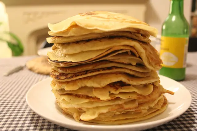 Talerz z naleśnikami na stole pokrytym obrusem w kratkę, za talerzem zielona butelka z napojem
