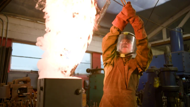 Ash em uma oficina. Ele usa um macacão de proteção e um capacete. Sai fogo de um recipiente no centro