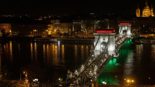 부다페스트의 체인 브리지가 야간에 헝가리 국기 색으로 밝혀졌고, 다리 위에 많은 사람들이 모여 있다.