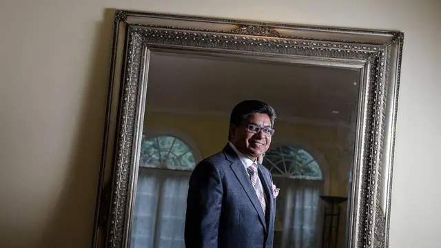 Una foto de Deepak Paradkar de 2013. Su reflejo es captado en un espejo con un marco plateado y ornamentado. Está mirando al espejo y sonriendo, lleva anteojos, un traje gris con una corbata rosa y un pañuelo de bolsillo a juego.