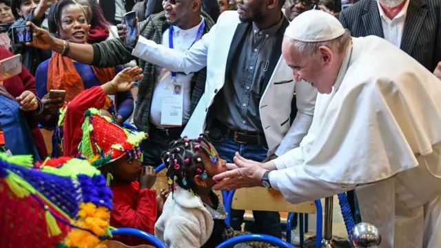 Le pape François en habits blancs tient le visage d'une jeune fille. La jeune fille porte une veste blanche et des cheveux tressés colorés. Un autre enfant portant un chapeau coloré et un haut rouge se tient derrière la fillette. Des personnes essaient de prendre des photos et des selfies avec le pape. 