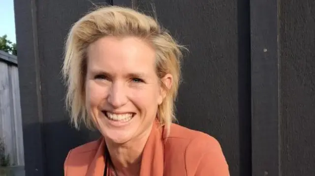 Una mujer de pelo rubio y con una chaqueta naranja sonría a cámara 