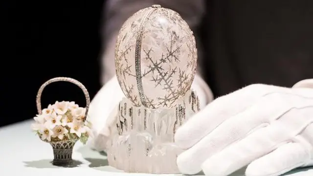 The Winter Egg is a clear egg with silver decoration on it in the shape of snowflakes. Next to it is an ornament of flowers in a basket. A white-gloved hand holds the egg while it sits on a white table.
