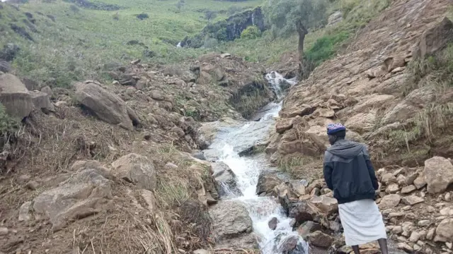 رجل يقف على حافة مجرى صخري في قرية ترسين بمنطقة جبل مَرَّة، ويرتدي جلابية بيضاء و سترة تجمع ما بين اللونين الأسود والرمادي