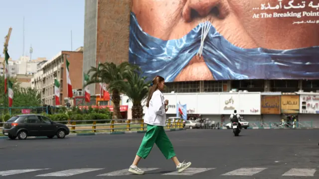 Uma mulher vestindo camisa branca e calça verde caminha em frente a um cartaz mostrando o Estreito de Ormuz como uma mordaça costurada na boca de Trump 