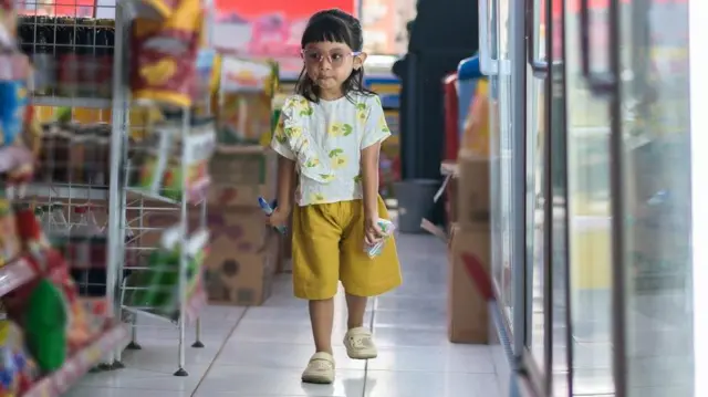 Seorang anak perempuan berjalan di dalam minimarket, memegang susu dan camilan sambil melihat-lihat rak. 