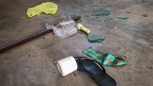 एक स्कूल के फ़र्श पर पड़े टूटे हुए कांच, चप्पल, टाई, डंडा आदि