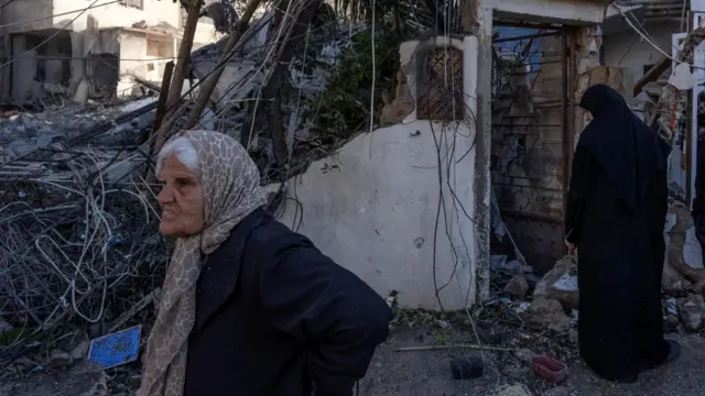 空爆により破壊された建物の前に、スカーフを頭からかぶった女性2人が立っている。建物はほとんどががれきになっているが、玄関の開口部だけ残っている