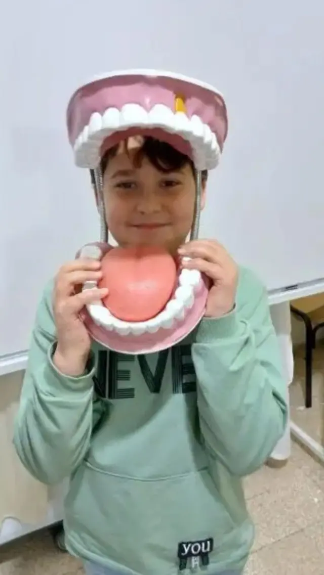 Kasım elinde ağız yapısını gösteren maket oyuncakla kameraya poz veriyor ve gülümsüyor.