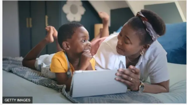 Aworan iya ati ọmọ rẹ kekere ti wọn n rẹrin-in lori ibusun pẹlu ẹrọ ayarabiaṣa lọwọ iya naa