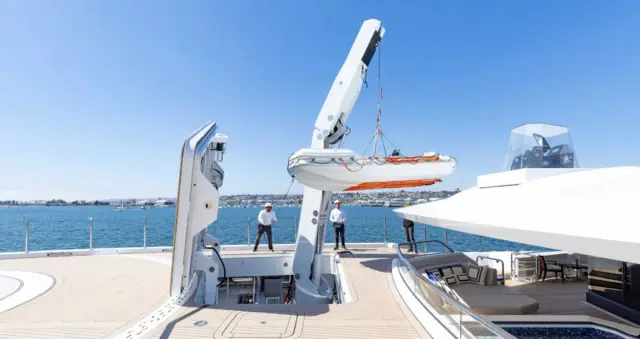 Güvertede denizci üniforması giymiş iki adam duruyor ve güvertenin üzerinde bir bot asılı. Helikopter pistinin kenarı sahnenin solunda, sağında ise beyaz örtülü bir oturma alanı var. Güneşli bir günde çekilmiş bir fotoğraf ve uzakta kara görünüyor.