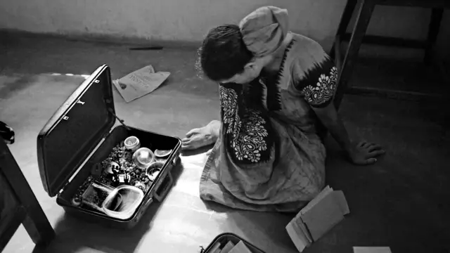 কন্যা শরণ্যার ব্যবহৃত জিনিসপত্র দেখে কান্নায় ভেঙে পড়া মা মিজ. সরোজিনী। ২০১৪ সালে, মাত্র ১৪ বছর বয়সে মারা যায় শরণ্যা। জন্ম থেকেই সে দৃষ্টিহীন ছিল, আবার হার্ট আর ফুসফুসের রোগও ধরা পড়েছিল। মিজ. সরোজিনীর স্বামীও ফুসফুসের রোগে মারা যান ২০০৬ সালে। এই পরিবারটি কাসারগড়ের একটি কাজু খেতের কাছে বাস করত