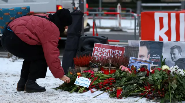 Человек в красной куртке, черной шапке, черных штанах и черных ботинках наклонился, чтобы положить цветы к мемориалу Навального.