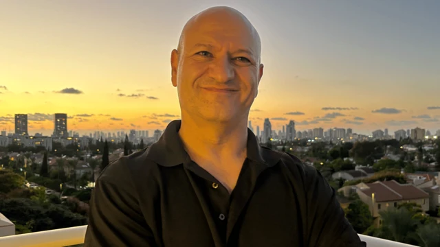 Michael Milshtein em uma sacada, sorri para a câmara. Ao fundo, o entardecer e os arranha-céus de uma cidade israelense